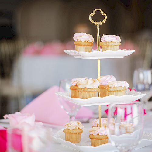 The image shows a tiered dessert stand with cupcakes on a decorated table, set with pink napkins, wine glasses, and flowers.