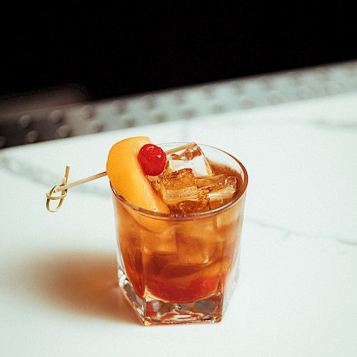 A glass of cocktail with ice cubes, garnished with an orange slice and a cherry on a skewer, placed on a white marble surface.