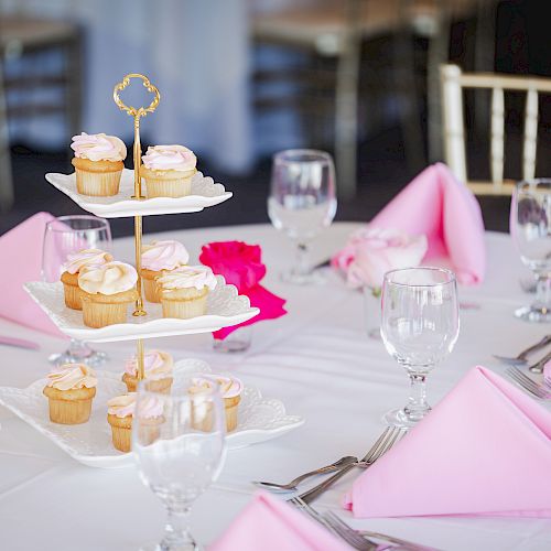 A table set for an event with tiered cupcakes, pink napkins, glassware, cutlery, and pink roses on a white tablecloth, creating an elegant atmosphere.