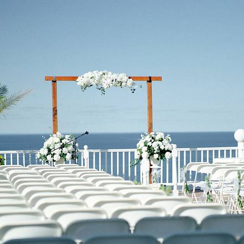 An outdoor wedding setup with white chairs, floral decorations, and a wooden arch facing the sea under a clear blue sky, creating a serene atmosphere.