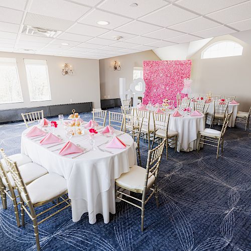 The image shows an elegantly decorated banquet room with round tables covered in white cloths, pink napkins, and gold chairs.