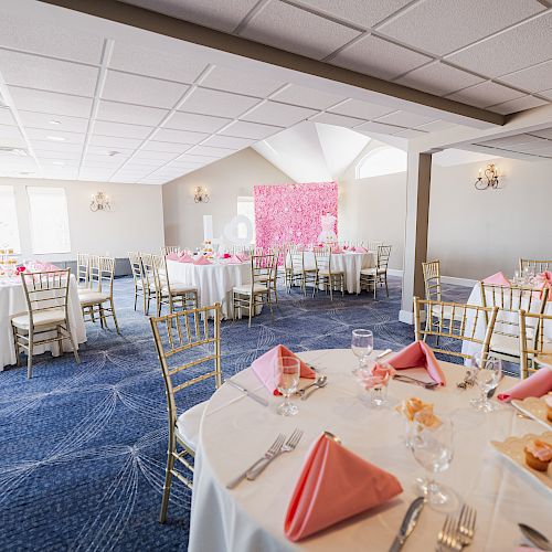 The image shows an elegantly set dining room with round tables, white tablecloths, pink napkins, and a cupcake stand, ready for an event.