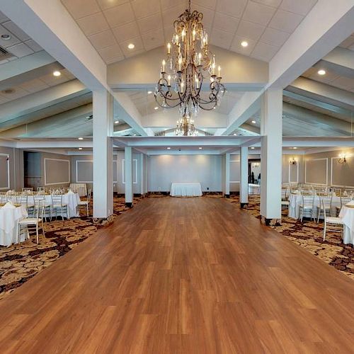 The image shows an elegant banquet hall with a wooden floor, chandeliers, white chairs, and tables set up on either side of the room.