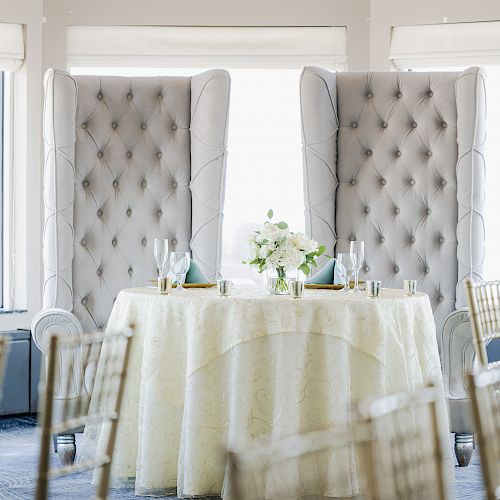 A beautifully set round table with a white cloth is adorned with flowers and flanked by two high-backed tufted chairs in a bright, elegant room.