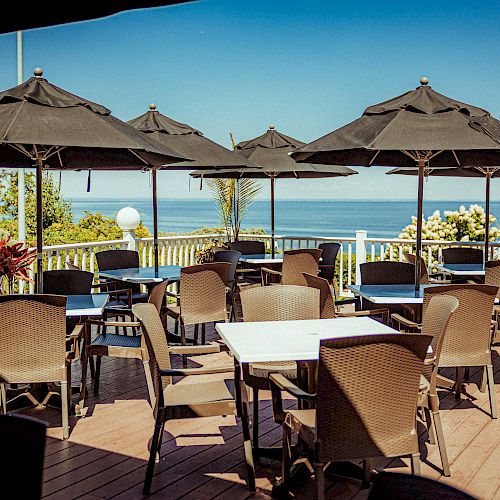 Outdoor patio with tables, chairs, and black umbrellas overlooking the ocean on a sunny day.