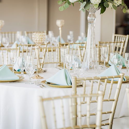 The image shows an elegantly decorated dining area with round tables set for an event, featuring light blue napkins, glassware, and gold chairs.