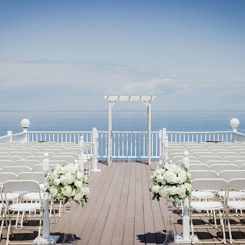 This image shows an outdoor wedding setup with rows of white chairs facing a white arch by the sea, under a clear blue sky, adorned with floral arrangements.