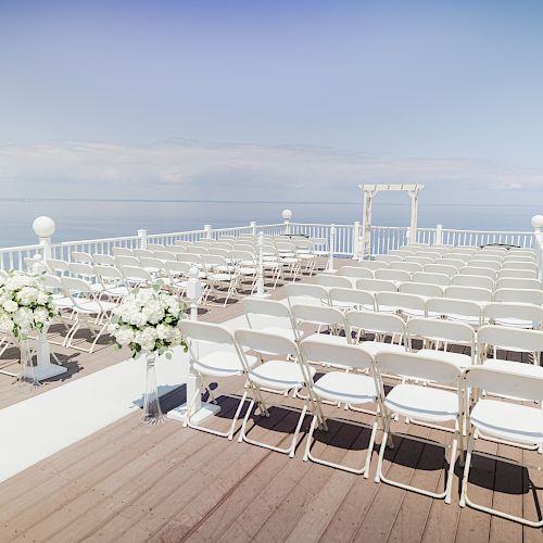 This image shows an outdoor wedding setup with white chairs arranged in rows, a floral arrangement on the left, and a small arch overlooking the ocean.