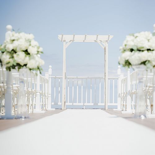 An outdoor wedding setup with white chairs and floral arrangements, leading to a gazebo under a clear blue sky.