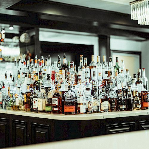 A bar counter filled with various bottles of liquor and spirits is shown, with a chandelier hanging overhead and part of the bar in the background.