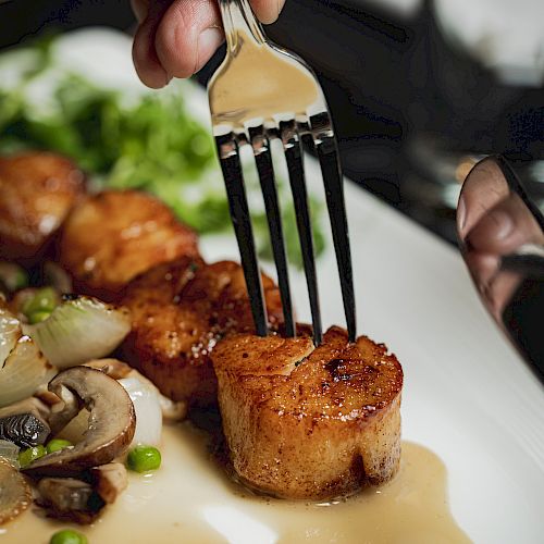 A person holding a fork with a seared scallop, alongside sautéed mushrooms and peas on a white plate.