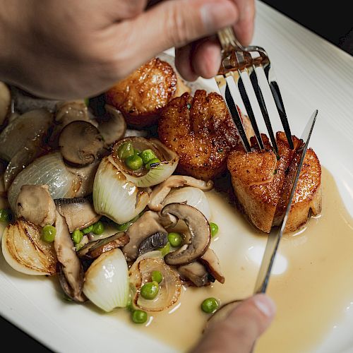 A person is cutting seared scallops on a plate with mushrooms, onions, peas, and sauce.