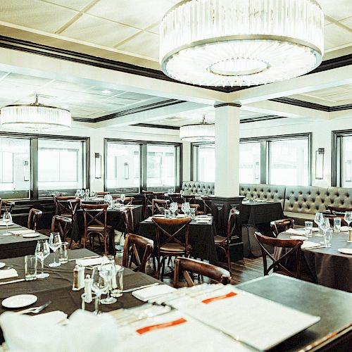 The image shows an elegant restaurant dining area with tables set with plates, glasses, and utensils, surrounded by chairs, featuring large windows and chandeliers.