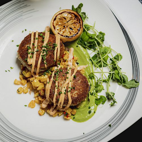 Two crab cakes on a bed of corn and salsa, garnished with sauce, microgreens, and a grilled lemon slice on a white plate.