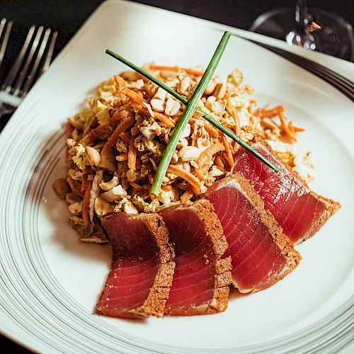 The image features a plate of seared tuna slices with a salad garnish, presented on a white dish, with utensils beside it on a dark background.