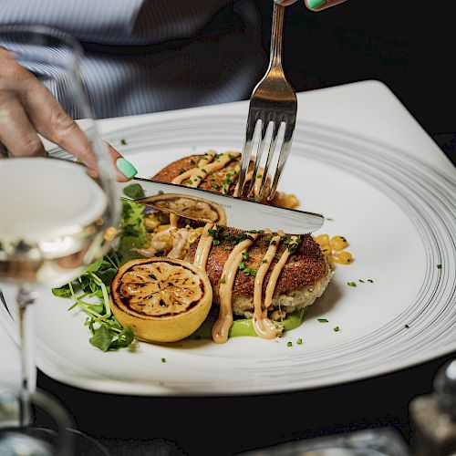 A person with green nails cuts a gourmet dish with a fork and knife next to a glass of white wine and a grilled lemon on a white plate.