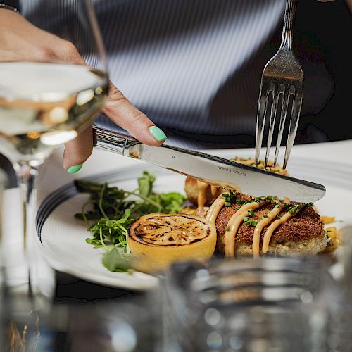 A person is enjoying a meal with a glass of white wine, greens, and a roasted lemon on a white plate.