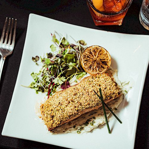 A plated meal with a breaded fish fillet, salad greens, a grilled lemon slice, and chives, shot from above with forks, a knife, and drinks visible.