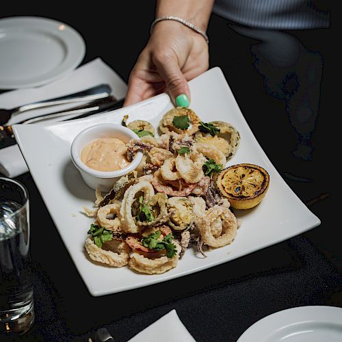 A hand holding a plated seafood dish garnished with herbs, a side of dipping sauce, a lemon half, over a table with utensils, plates, and a water glass.