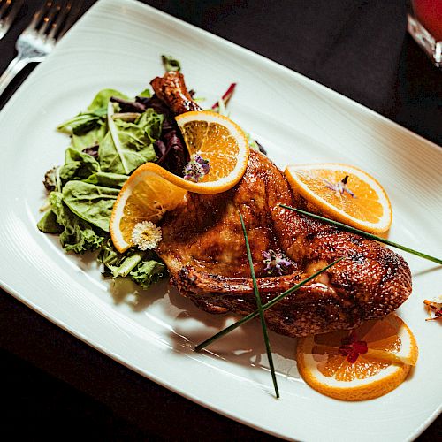 It's a plated roasted chicken garnished with orange slices, served with leafy greens and accompanied by utensils on a table.