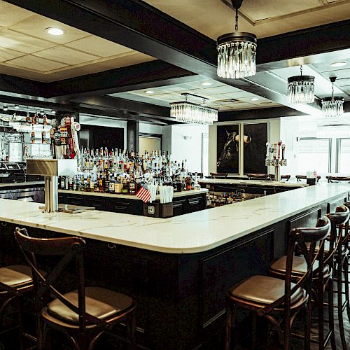 An elegant bar with a large, marble-topped counter, wooden chairs, and well-stocked shelves of liquor bottles under chandeliers and natural lighting.