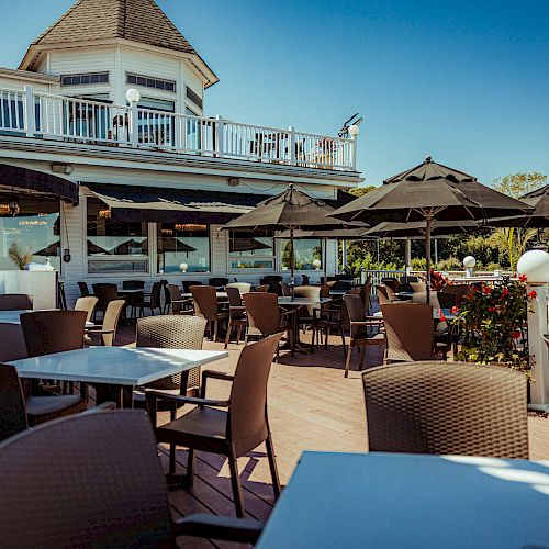 An outdoor restaurant seating area with tables, chairs, and black umbrellas, adjacent to a two-story building with white railings, in daylight.