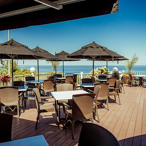 An outdoor restaurant patio with dark umbrellas over tables and chairs, overlooking a clear blue ocean under a sunny sky.