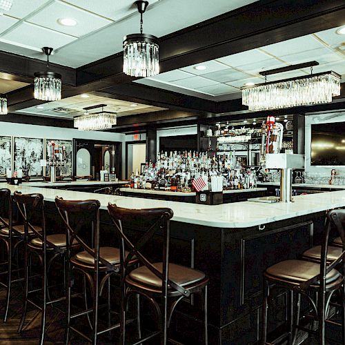 A spacious, modern bar with high chairs, chandeliers, and a well-stocked counter. The atmosphere is sophisticated and inviting.