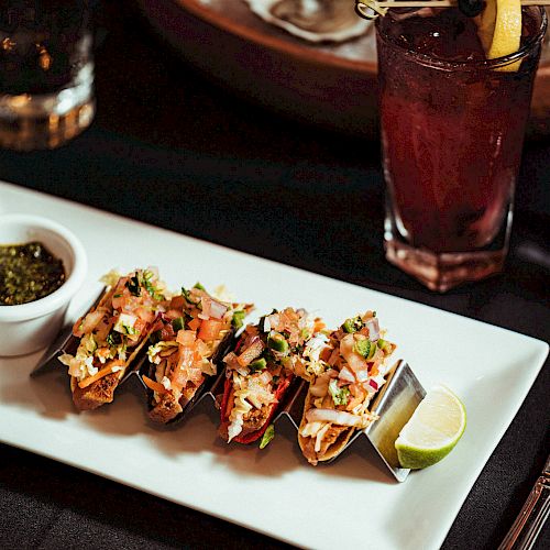 The image shows a plate with four tacos, a sauce dip, and a lime wedge on a table with a dark drink garnished with a lemon slice and oysters in the background.