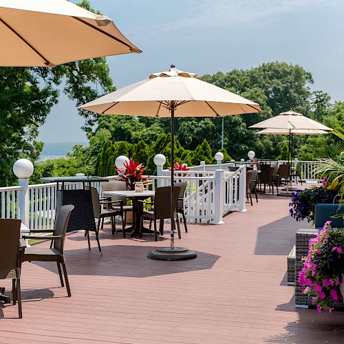 An outdoor deck with tables, chairs, umbrellas, and potted plants, set against a backdrop of trees and a distant body of water.