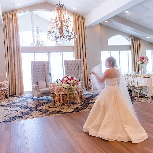 A bride in a white dress stands on a wooden dance floor in an elegant reception hall with chandeliers, floral arrangements, and table settings.