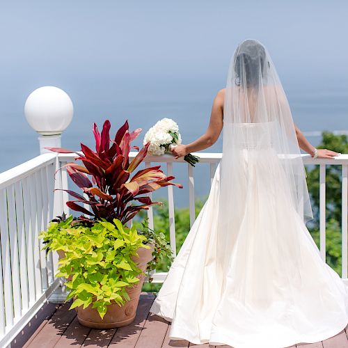 A bride in a wedding dress and veil stands on a balcony, holding a bouquet, overlooking a scenic view with greenery and the ocean.