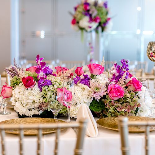 The image shows a beautifully set dining table with a centerpiece of pink, white, and purple flowers, surrounded by golden candle holders and glasses.