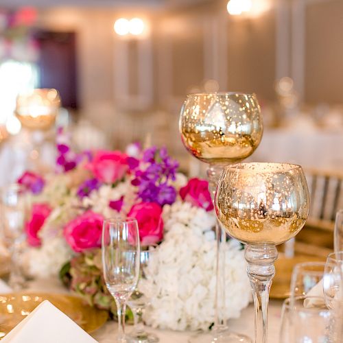 The image shows a beautifully set table with golden decorative glasses, colorful flowers, and elegant tableware at an event venue.