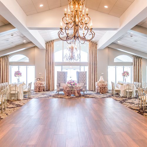 The image shows a large, elegant banquet hall with chandeliers, decorated tables, chairs, flowers, and large windows providing natural light.