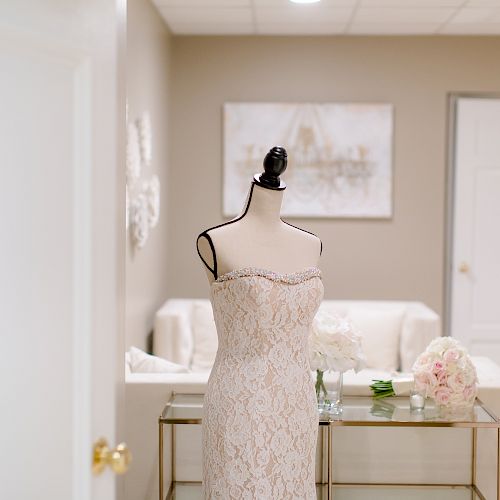 The image shows a wedding dress on a mannequin in a well-lit room with neutral walls, a door, a painting, and stylish furniture.