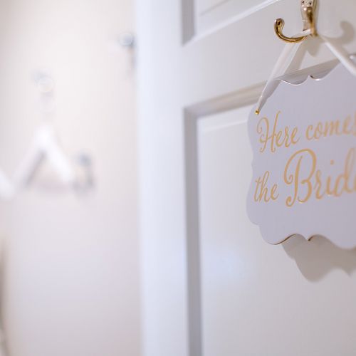 A sign reading "Here comes the Bride" hangs on a door, with blurred white objects, possibly wedding attire, visible in the background.