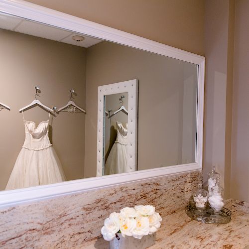 A white wedding dress hangs on a wall, reflected in a large vanity mirror; the countertop holds white flowers and glass containers filled with decorative items.