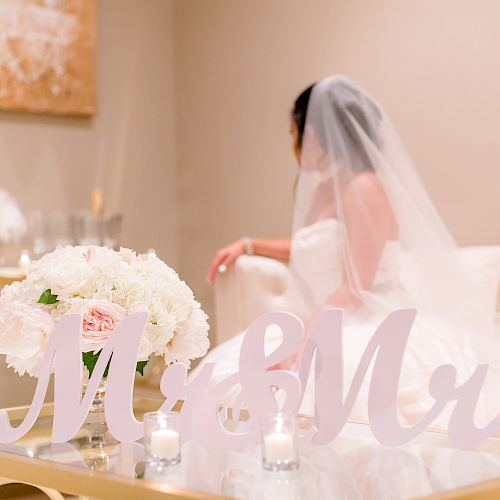 A bride in a veil sits on a couch with a "Mr & Mrs" sign and white flowers in front of her, along with candles on a glass table.