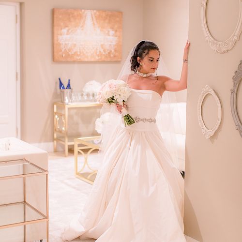 A bride in a white wedding dress holds a bouquet, posing in a decorated room with mirrors and furniture.