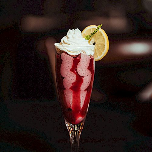 The image shows a tall glass of a pink or red beverage topped with whipped cream and garnished with a lemon slice, set against a dark background.