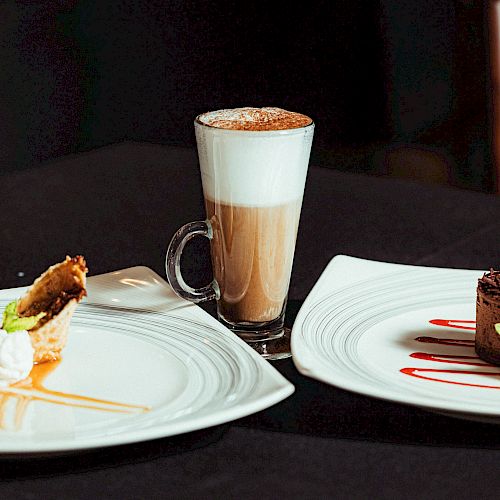 A cappuccino with foam is served alongside a dessert plate with two pastries and another with a chocolate dessert, each garnished with cream.