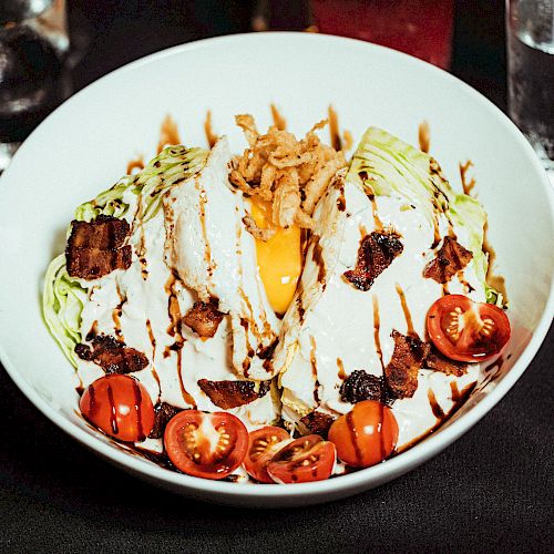 A bowl of salad featuring lettuce, cherry tomatoes, bacon bits, a poached egg, and creamy dressing, garnished with crispy onions and a sauce drizzle.