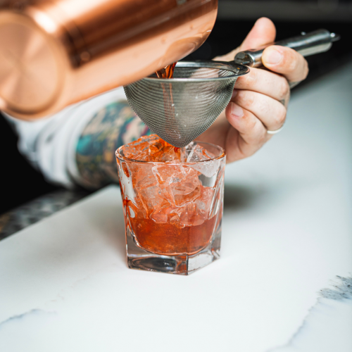 A bartender is pouring a red cocktail into a glass with ice cubes, using a mesh strainer for precision.