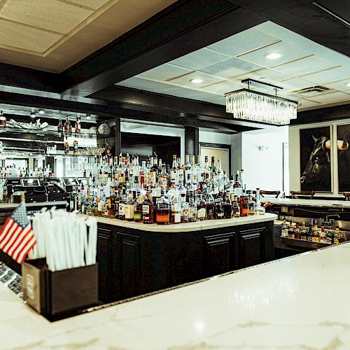 A well-stocked bar with various bottles, a U.S. flag, and a straw holder. The setting is modern with ambient lighting and a bull painting on the wall.