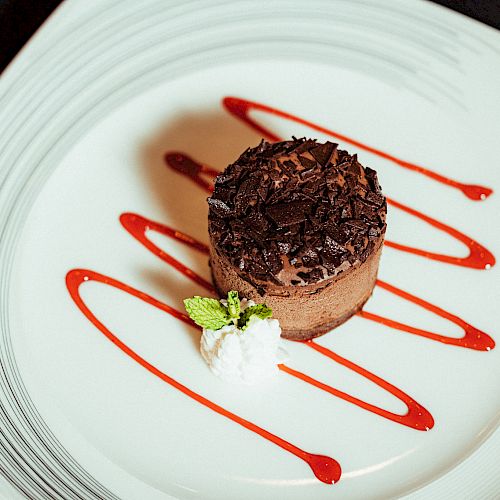 A chocolate dessert garnished with chocolate shavings, served with raspberry sauce and a dollop of cream with a mint leaf, on a white plate.