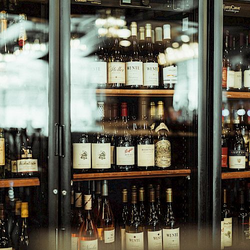 The image displays a variety of wine bottles stored in a glass-door cooler at a wine shop or bar.