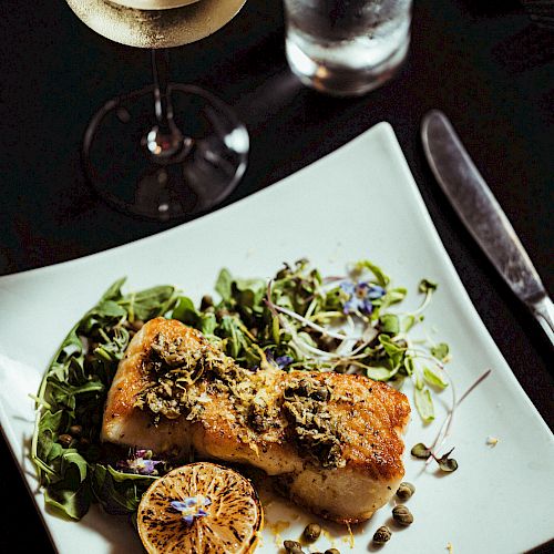 A beautifully plated fish dish garnished with greens and a lemon half, accompanied by a glass of white wine and a glass of water on a dark table.