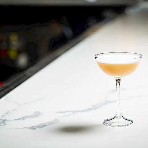 A cocktail in a coupe glass sits on a marble countertop with a blurred bar background.