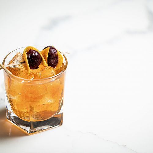 A glass of an amber-colored cocktail with ice, garnished with two dark cherries on a skewer. The glass is placed on a white marble surface.